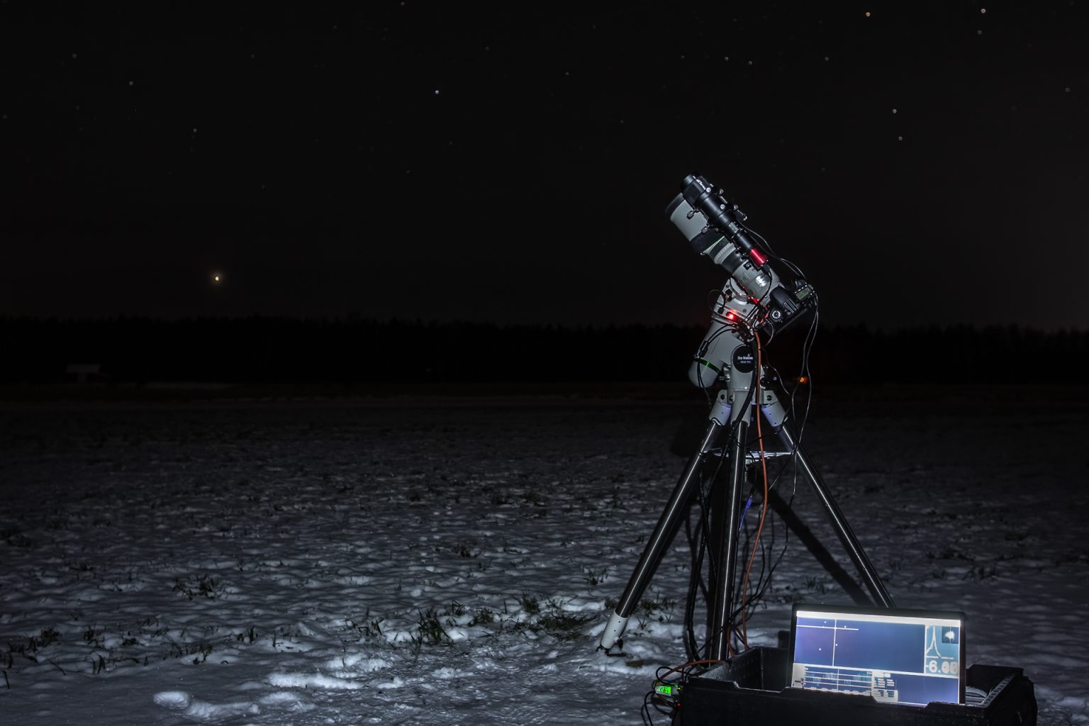 Astrokuvaus meneillään. Kamera seisoo jalustalla lumihangessa pimeällä pellolla. Jannella on käytössään JAS Kamerahuollossa astromodattu  Canon EOS 60D kamera, Canon EF 400mm f/4 DO IS II USM objektiivi ja Heq-5 pro synscan seurantajalusta.